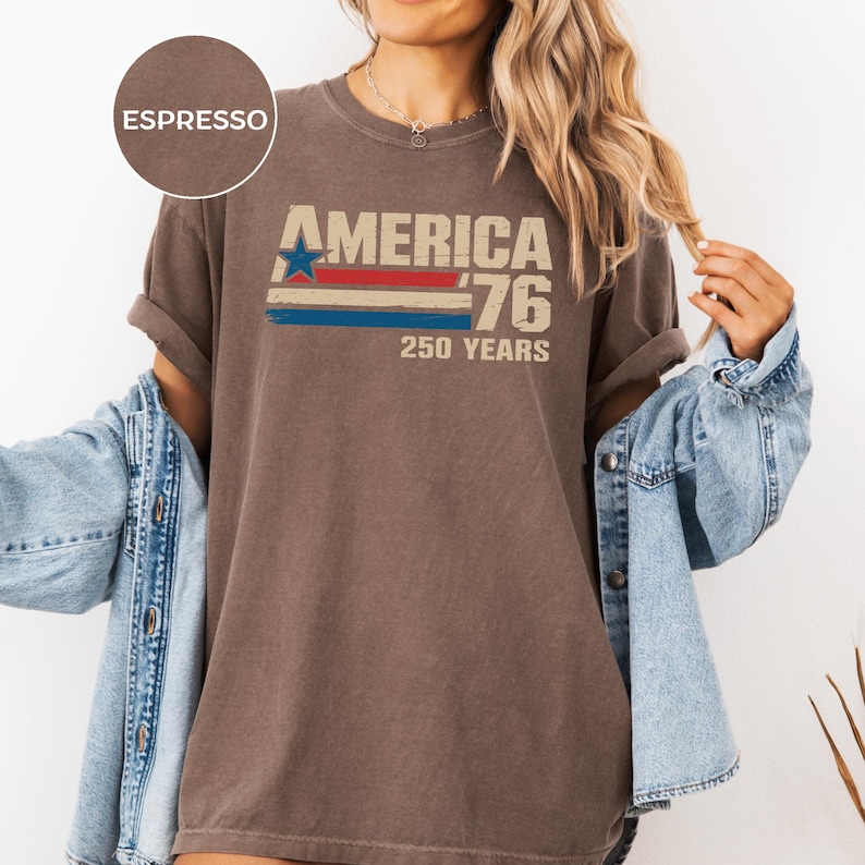 May include: Espresso-coloured t-shirt featuring the text "AMERICA '76 250 YEARS" in a vintage style. The design includes a blue star and red and blue stripes. The shirt has rolled-up sleeves and is paired with a denim jacket.