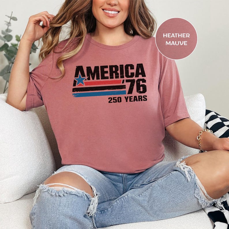 May include: Mauve t-shirt with the text "AMERICA '76 250 YEARS" in black, red, blue, and white. The shirt features a star graphic and horizontal stripes. The shirt is worn with ripped blue jeans.