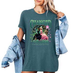 K&ouml;nnte beinhalten: T&uuml;rkisfarbenes T-Shirt mit einer Grafik von zwei Hexen und dem Text "CHANGED FOR GOOD". Das Design zeigt eine gr&uuml;ne Hexe in einem schwarzen Kleid und eine rosa gekleidete Hexe. Text und Design sind in Gr&uuml;n- und Rosat&ouml;nen gehalten.