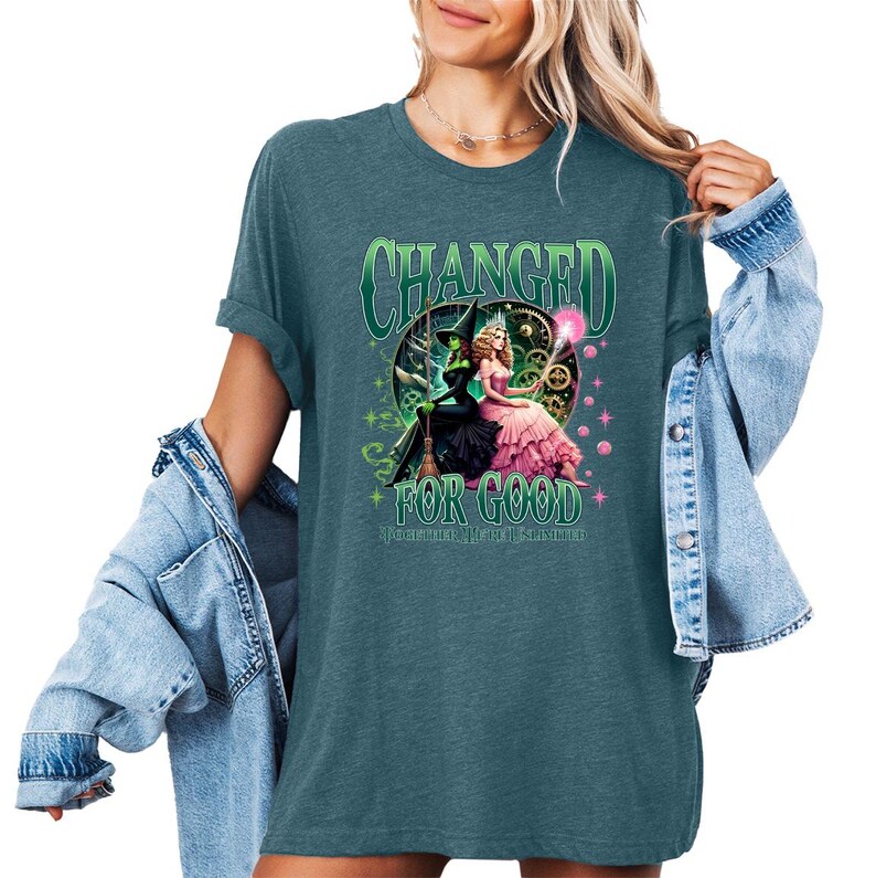 K&ouml;nnte beinhalten: T&uuml;rkisfarbenes T-Shirt mit einer Grafik von zwei Hexen und dem Text "CHANGED FOR GOOD". Das Design zeigt eine gr&uuml;ne Hexe in einem schwarzen Kleid und eine rosa gekleidete Hexe. Text und Design sind in Gr&uuml;n- und Rosat&ouml;nen gehalten.