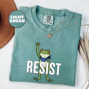 May include: Light green t-shirt with a cartoon frog graphic. The frog is raising a fist and wearing a blue bandana. The word "RESIST" is printed in large white letters below the frog. A silver necklace is visible.