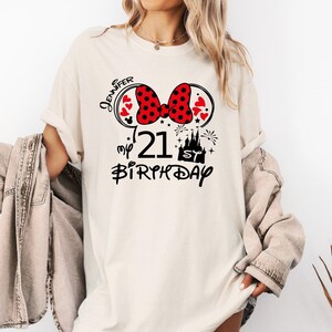 May include: Cream-colored t-shirt with a graphic design for a 21st birthday. The design includes a red polka-dot bow, Mickey Mouse ears, hearts, a castle, fireworks, and the text "Jennifer", "21", and "Birthday".