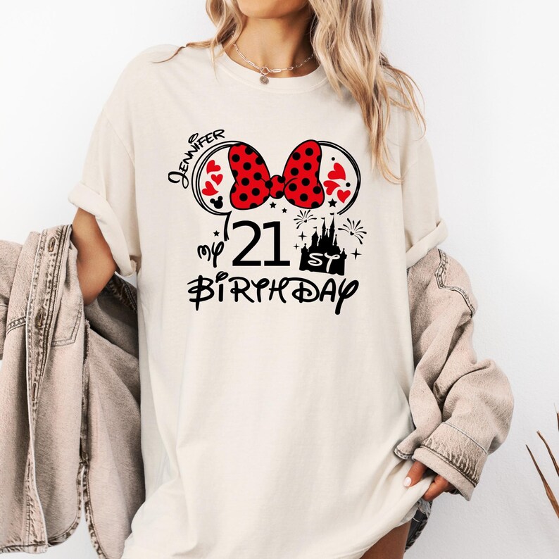 May include: Cream-colored t-shirt with a graphic design for a 21st birthday. The design includes a red polka-dot bow, Mickey Mouse ears, hearts, a castle, fireworks, and the text "Jennifer", "21", and "Birthday".
