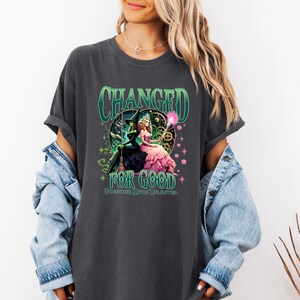 K&ouml;nnte beinhalten: Dunkelgraues T-Shirt mit einem Motiv aus dem Musical "Wicked". Das Design zeigt den Text "CHANGED FOR GOOD" in Gr&uuml;n, mit Illustrationen von Elphaba und Glinda. Das Shirt ist ein bequemer, l&auml;ssiger Stil.