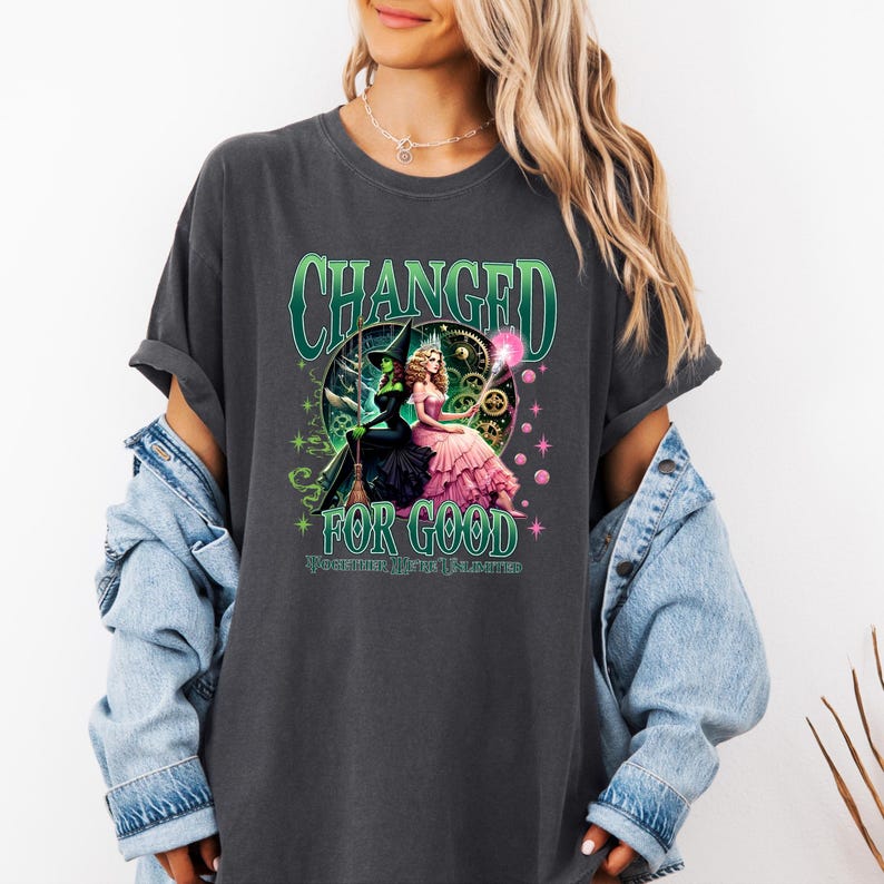 K&ouml;nnte beinhalten: Dunkelgraues T-Shirt mit einem Motiv aus dem Musical "Wicked". Das Design zeigt den Text "CHANGED FOR GOOD" in Gr&uuml;n, mit Illustrationen von Elphaba und Glinda. Das Shirt ist ein bequemer, l&auml;ssiger Stil.