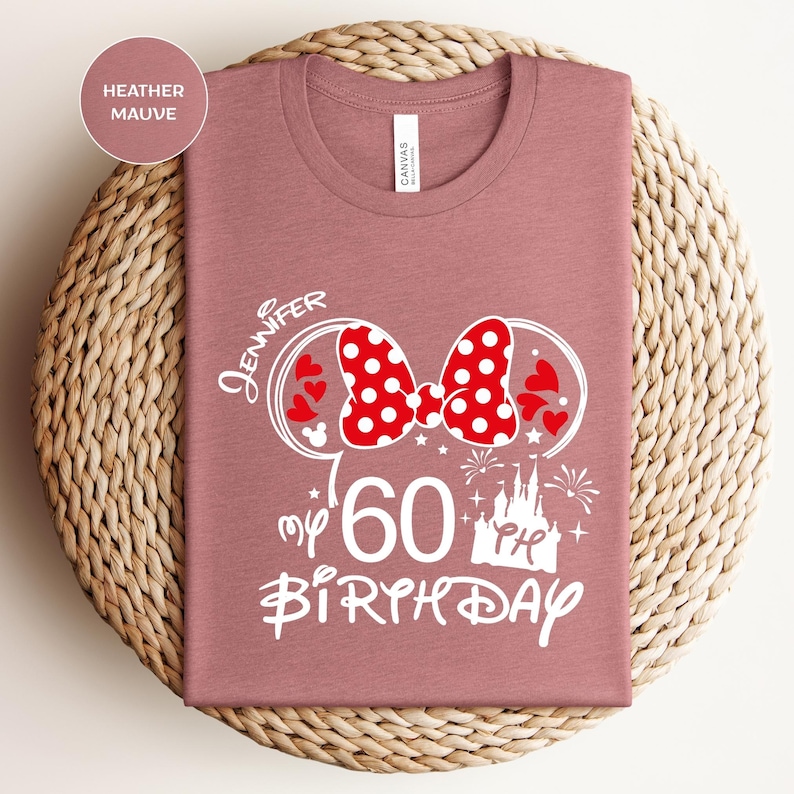 Puede incluir: Camiseta color malva con un dise&ntilde;o gr&aacute;fico blanco. El dise&ntilde;o incluye un lazo rojo de lunares, orejas de Mickey Mouse, corazones, un castillo, fuegos artificiales y el texto "Jennifer my 60th Birthday". La camiseta est&aacute; doblada sobre una cesta tejida.