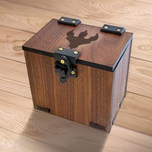 May include: A wooden box with a dark brown finish, featuring a decorative emblem on the lid. The box has black metal hinges and a latch. The box is sitting on a wooden surface.