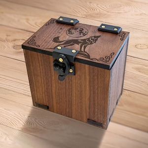 Op de afbeelding: Een vierkante houten kist met een ontwerp van een huilende wolf en maan op het deksel. De kist is warmbruin gebeitst en heeft zwarte metalen beslag en decoratieve hoekaccenten. Het heeft een sluiting met een grendel.