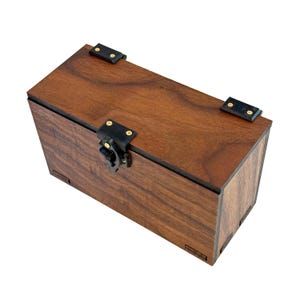 May include: A rectangular wooden box with a dark brown finish. The box has a hinged lid and is secured with a black clasp and hinges. The box is made of wood and has a natural wood grain pattern.