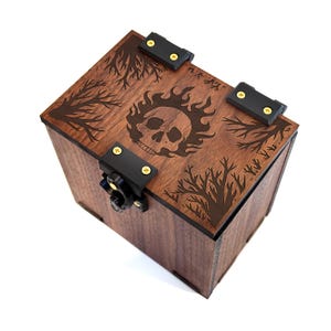 Puede incluir: Una caja de madera marrón con un diseño de calavera y llamas en la tapa. La caja tiene herrajes negros y un pestillo. La tapa también presenta diseños de ramas de árboles en las esquinas.