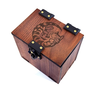 May include: A wooden box with a dark brown finish, featuring a laser-etched design of a cat curled in a circle on the lid. The box has black metal hinges and a clasp. The box is a cube shape.
