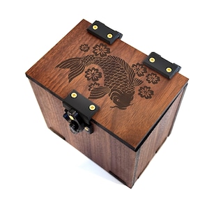 May include: A wooden box with a dark brown finish, featuring a laser-etched koi fish and floral design on the lid. The box has black metal hinges and a clasp with gold-colored screws. The box is square-shaped.