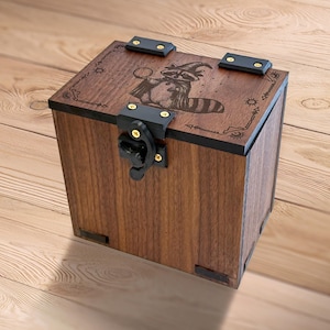 May include: A wooden box with a raccoon wizard design etched on the lid. The box is brown with black metal accents and a latch. The raccoon is wearing a wizard hat and cape, holding a glowing orb.