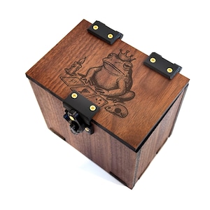 May include: A dark brown wooden box with a black clasp and hinges. The top of the box features an engraved illustration of a frog wearing a crown and playing cards. The box is square and has a decorative design.