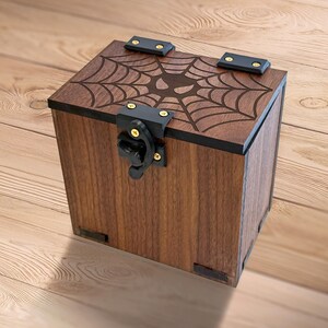 Könnte beinhalten: Eine dunkelbraune Holzkiste mit einem eingravierten Spinnennetz-Design auf dem Deckel. Die Kiste hat schwarze Metallscharniere und einen Verschluss. Die würfelförmige Kiste hat ein handwerkliches Aussehen mit rustikaler Ästhetik.
