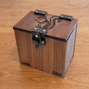 May include: A wooden box with a squirrel engraving on the lid. The box is made of dark brown wood with black accents and a black latch. The box is square-shaped and appears to be handcrafted.