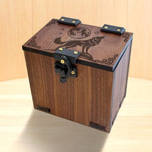 Op de afbeelding: Een vierkante houten doos met een donkerbruine afwerking, voorzien van een lasergegraveerd ontwerp van een wolf die naar een volle maan huilt op het deksel. De doos heeft zwarte metalen scharnieren en een bijpassende zwarte sluiting met goudkleurige schroeven.