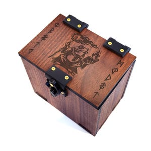 Puede incluir: Una caja de madera marrón oscuro con un diseño grabado con láser de un mago rana sosteniendo un bastón y un libro. La caja tiene bisagras y cierres de metal negro con tornillos dorados. Símbolos decorativos grabados en los bordes.