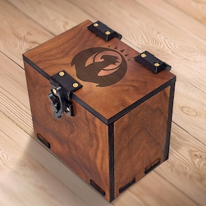 Könnte beinhalten: Eine Holzbox mit dunkler Beize und einem dekorativen Emblem auf dem Deckel. Die Box hat schwarze Scharniere und einen Verschluss. Die quadratische Box besteht aus mehreren Holzplatten. Die Box steht auf einer Holzoberfläche. Die Box ist etwa 15 cm hoch.