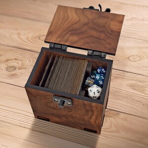 Könnte beinhalten: Eine dunkelbraune Holzbox mit Klappdeckel, die einen Stapel rechteckiger Karten und mehrere mehrseitige Spielwürfel enthält. Die Box verfügt über einen schwarzen Metallverschluss an der Vorderseite und schwarze Metallscharniere am Deckel.