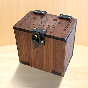 May include: A dark brown wooden box with a hinged lid featuring a laser-engraved frog, moon phases, and geometric symbols. The box includes black metal hinges and a black metal latch with gold-coloured screws, providing a secure closure mechanism.