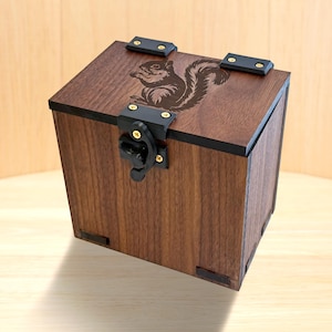 May include: A square wooden box with a dark brown finish featuring a laser-engraved squirrel on the lid. The box includes black metal hinges and a black metal latch mechanism secured with small gold-colored screws on the front and top panels.