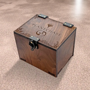 May include: A wooden box with the word "PLANECHASE" and a logo engraved on the lid. The box is dark brown with black metal hinges and a clasp. The box is square and appears to be handmade.