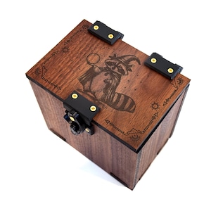 May include: A wooden box with a dark brown finish, featuring a laser-etched design of a raccoon wearing a wizard hat and cape. The box has black metal clasps and hinges with gold-colored screws. The design includes sun and star motifs.