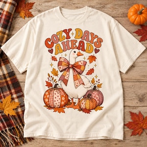 May include: A cream-colored t-shirt featuring a graphic of a white goose wearing a large plaid bow, surrounded by three patterned pumpkins and autumn leaves. The text "COZY DAYS AHEAD" is printed in a retro orange and yellow font above the goose.
