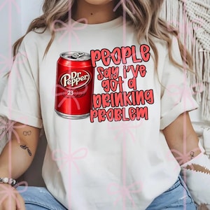 May include: Cream-colored t-shirt featuring a red Dr. Pepper can graphic with the text "People Say I've Got a Drinking Problem" in pink and black. The shirt has short sleeves and a relaxed fit.