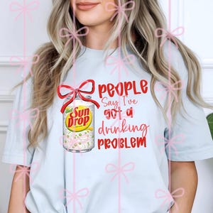 May include: Light blue t-shirt featuring a Diet Sun Drop soda can with a red bow and the text "People Say I've Got a Drinking Problem" in red. The soda can has a yellow and green label.