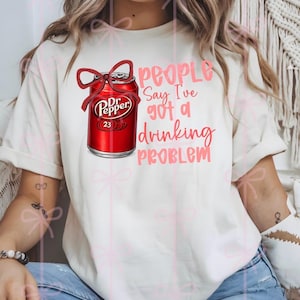 May include: Cream-colored t-shirt featuring a red Dr. Pepper can with a bow, and the text "People Say I've Got a Drinking Problem" in pink. The shirt is a casual style.
