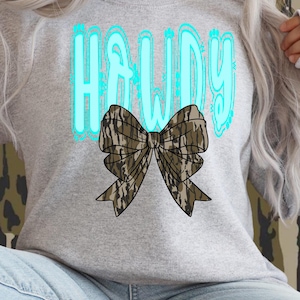 May include: A gray sweatshirt with the word "HOWDY" in turquoise letters. Below the text is a large bow with a camouflage pattern. The sweatshirt is a casual style.