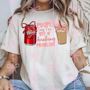 Könnte beinhalten: Cremefarbenes T-Shirt mit einer Grafik einer Dr. Pepper-Dose und einem Eiskaffee, beide mit Schleifen verziert. Der rosa Text lautet: „People Say I've Got a Drinking Problem“.