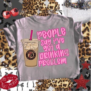 May include: A light gray t-shirt with the phrase "People say I've got a drinking problem" in pink lettering. The shirt features an illustration of an iced coffee drink with the "Seven Brew" logo.