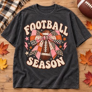 May include: A charcoal gray short-sleeve t-shirt featuring a graphic print of a brown football tied with a large, multi-patterned pink bow. The text "FOOTBALL SEASON" is printed in a cream-colored retro font, accented by small pink lightning bolts and stars.