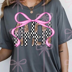 May include: Gray t-shirt with the word "AMEN" in a bold, decorative font. The letters feature a mix of leopard print and black and white checkerboard patterns, accented by a pink bow. A casual, comfortable style.