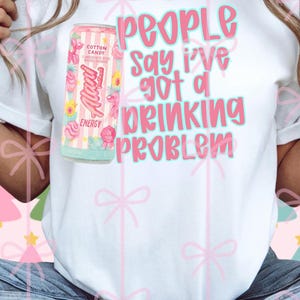 Op de afbeelding: Wit T-shirt met de tekst "People say I've got a drinking problem" in roze en blauw. Een blikje "Alani Nu" Energydrink met suikersmaak staat naast de tekst. Het blikje is roze met bloemmotieven.