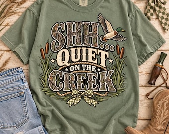 May include: A sage green short-sleeve t-shirt featuring a graphic print with the text SHH QUIET ON THE CREEK. The design includes a flying mallard duck, cattails, and a checkered bow. The letters have a camouflage pattern and rope border.
