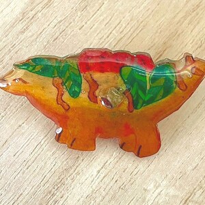 Könnte beinhalten: Eine farbenfrohe Brosche aus Harz in Form eines stilisierten Tieres mit grünen, roten und orangefarbenen Details. Die Brosche hat eine glänzende Oberfläche und ein verspieltes Design.