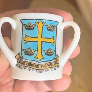 Puede incluir: Una pequeña taza de cerámica blanca con dos asas. La taza presenta un escudo azul y dorado con una cruz y coronas. El texto en la taza dice "King Edward the Martyr Assassinated at Corfe Castle AD 978."