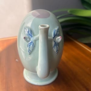 May include: A pale green ceramic teapot with a rounded body and a spout. The teapot has blue leaf and pink bud accents. The lid is a darker shade of green with a purple centre. The teapot sits on a small, round base.