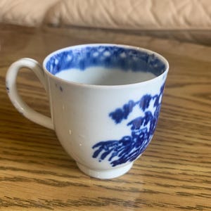 Puede incluir: Una taza de té de porcelana blanca con un diseño floral azul y blanco. La taza tiene un asa y una base redondeada.