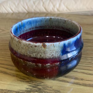 Puede incluir: Un pequeño cuenco redondo de cerámica con un esmalte azul, blanco y rojo. El cuenco tiene un borde ligeramente irregular y una superficie texturizada.