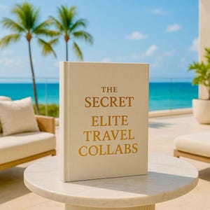 May include: A cream-coloured hardcover book with the title "THE SECRET ELITE TRAVEL COLLABS" in gold lettering. The book rests on a white marble table. The background shows palm trees, a blue ocean, and a clear sky.