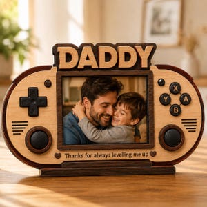 Op de afbeelding: Houten fotolijst in de vorm van een gamecontroller met het woord "DADDY" bovenaan. De lijst bevat een foto van een vader en kind. De tekst "Thanks for always levelling me up" staat onderaan. De lijst is van licht en donker hout.