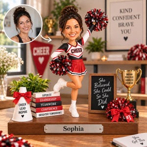 Può includere: Una statuetta bobblehead personalizzata di una cheerleader con capelli castani raccolti in uno chignon, che indossa un'uniforme nera, bianca e rossa. La figura tiene pompon rossi, bianchi e neri, poggia su una base in legno con l'etichetta Sophia ed è circondata da libri in miniatura, un trofeo e un cartello con la scritta She Believed She Could So She Did.