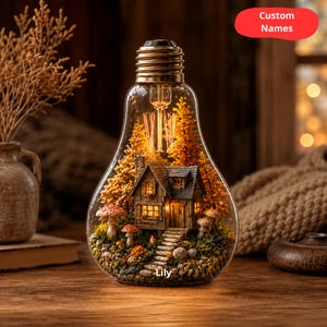 May include: A decorative light bulb containing a miniature woodland scene. The scene features a small house, trees, and mushrooms. The text "Custom Names" is on a red oval, and the name "Lily" is at the bottom.