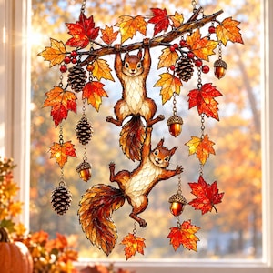 May include: A stained glass suncatcher featuring two brown squirrels hanging from a tree branch adorned with vibrant red and orange maple leaves, small pinecones, and acorns, all suspended by delicate metal chains against a window.