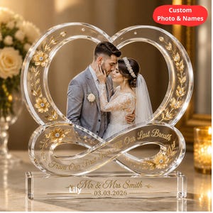 Peut inclure: Cadre photo en forme de cœur en acrylique transparent avec des détails floraux dorés et l'inscription "From Our First Kiss Till Our Last Breath". Le cadre contient une photo de couple. La base porte un nom et une date.